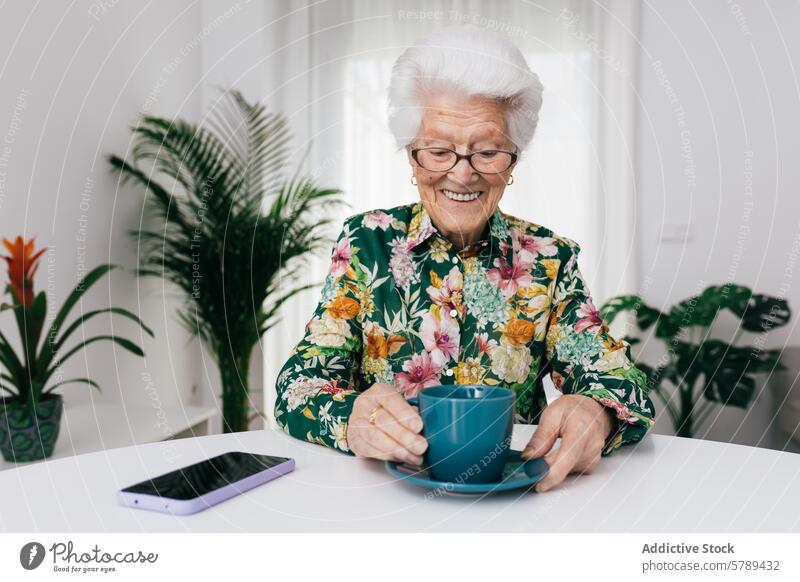 Ältere Frau genießt eine Tasse Tee zu Hause Senior älter heimwärts Freude geblümt Bluse Fröhlichkeit weiße Haare Lächeln Brille Zimmerpflanze Raum heimisch