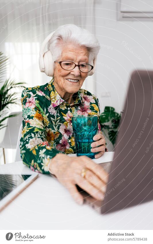 Ältere Frau, die zu Hause mit Kopfhörern am Laptop arbeitet Senior Technik & Technologie Lächeln älter aktives Altern heimwärts arbeiten Blumenhemd Brille
