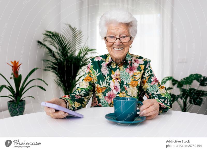 Ältere Frau lächelt zu Hause mit Kaffee und Smartphone älter Senior Lächeln heimwärts Brille geblümt Hemd Tasse trinken Beteiligung Tisch weiß Zimmerpflanze