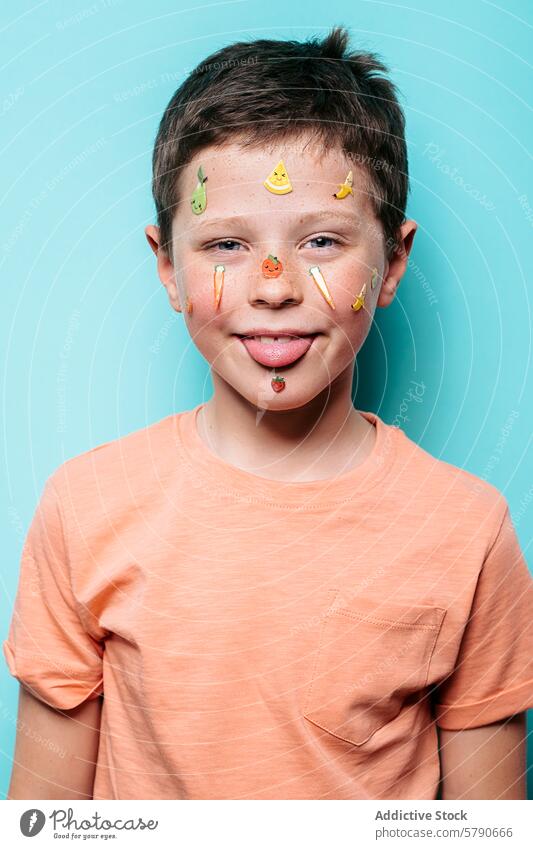 Fröhlicher Junge mit fruchtigen Aufklebern im Gesicht und herausgestreckter Zunge Kind heiter Freude spielerisch Frucht farbenfroh türkis Hintergrund jugendlich