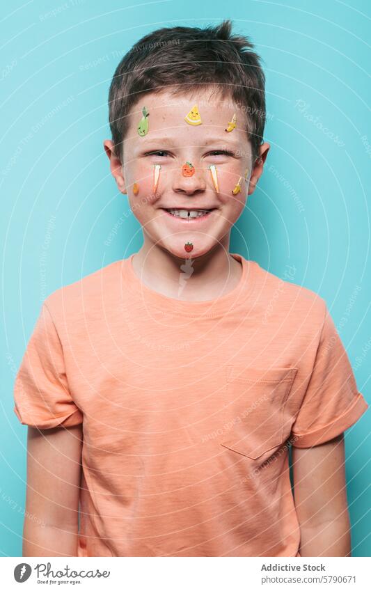Fröhlicher Junge mit Obstaufklebern im Gesicht und türkisfarbenem Hintergrund Lächeln freudig Frucht Aufkleber blaugrün Blauer Hintergrund Zierde farbenfroh
