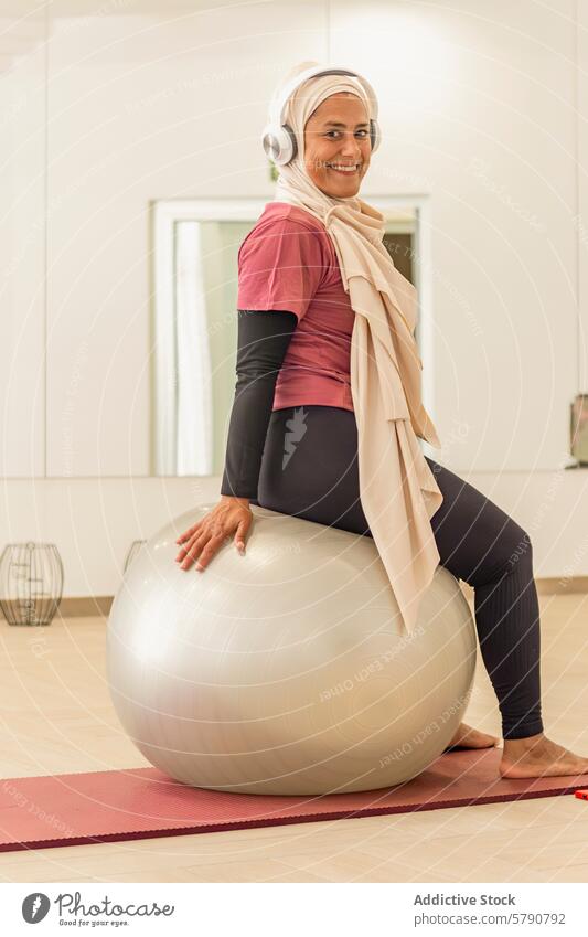 Muslimische Frau lächelnd auf einem Fitnessball vor einer Yogastunde muslimisch Fitnessstudio Training Kopfhörer Vorbereitung Klasse aktiv Lifestyle Gesundheit