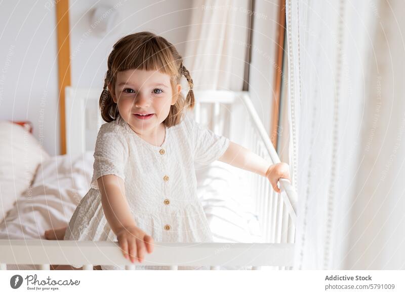Entzückendes Kleinkind im weißen Kleid an der Krippe stehend Mädchen weißes Kleid Babybett Schlafzimmer Kind Lächeln Zopf sonnig heiter Unschuld niedlich