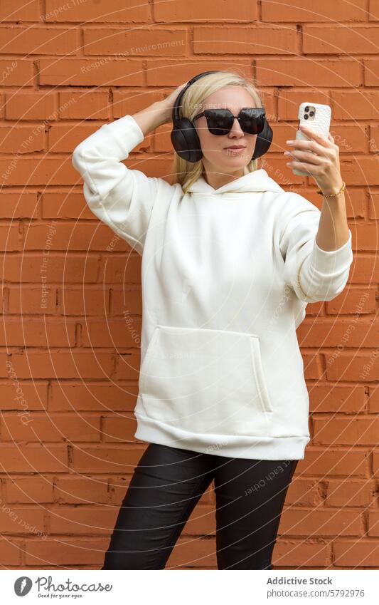 Stilvolle Frau mit Smartphone und Kopfhörern auf einer Straße in der Stadt blond Musik lässig Kleidung Backsteinwand urban Mode Sonnenbrille Selfie