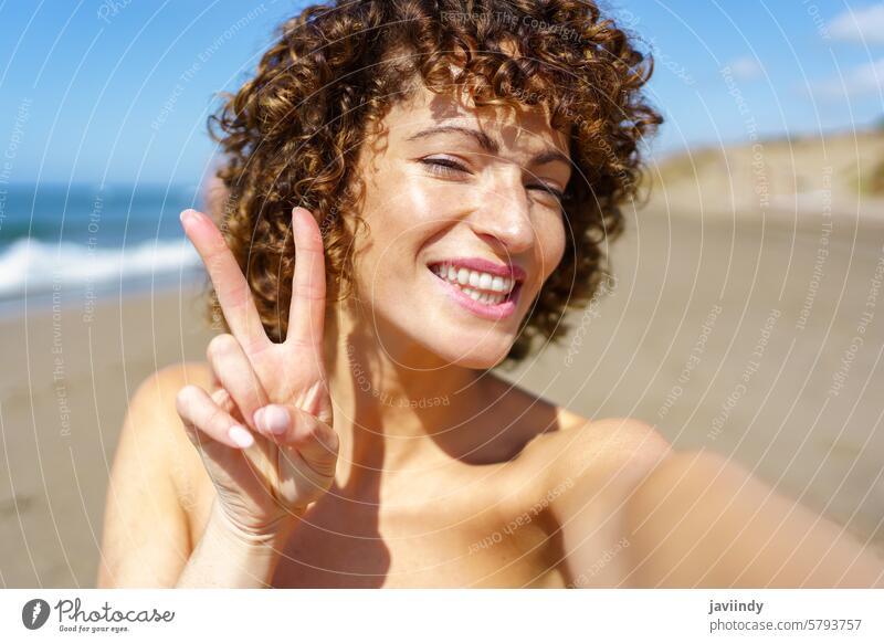 Glückliche Frau mit V-Zeichen am Strand Selfie Urlaub Lächeln Sommer v-Zeichen zwei Finger gestikulieren heiter jung krause Haare Moment positiv Meer Kälte MEER
