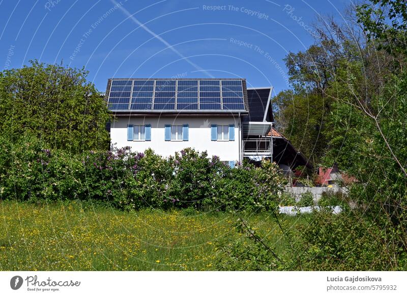 Auf dem Dach eines weißen Einfamilienhauses installierte Solar- oder Fotovoltaikmodule, beobachtet an einem sonnigen Tag in einem Schweizer Dorf. Um das Haus herum stehen Bäume und Büsche.