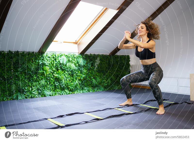 Lächelnde Frau in der Hocke im Fitnessstudio beim Training mit der Agility-Leiter Athlet Kniebeuge springen agil Gesundheit Übung Sportlerin jung krause Haare
