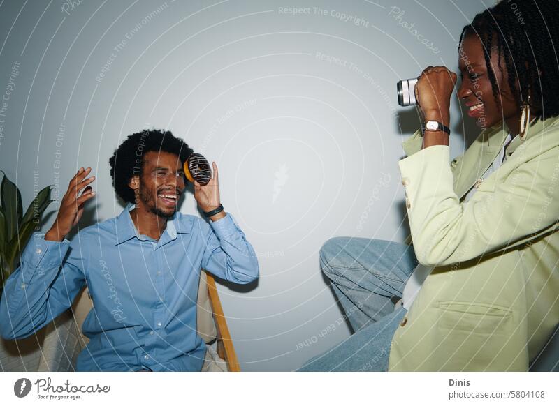 Smiling creative Black woman photographing coworker in office posing fun party donut playful happy celebrate friday businesswoman colleague Black man