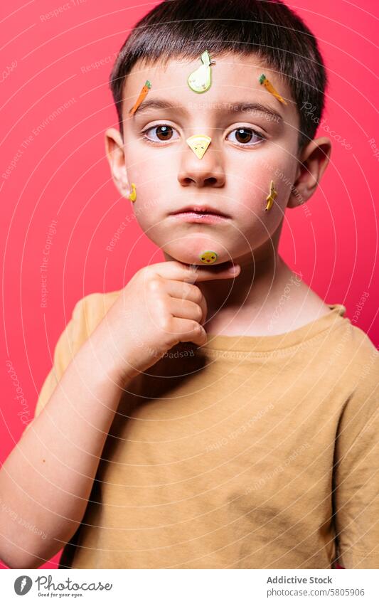 Junge mit Fruchtaufklebern im Gesicht auf rosa Hintergrund Kind Aufkleber Klebstoff Porträt beschaulich in die Kamera schauen expressiv Schulkind spielerisch