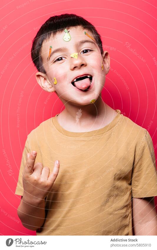 Verspielter Junge mit Aufklebern im Gesicht macht dummes Gesicht spielerisch Zunge gestikulieren rosa Hintergrund heiter jung Kind Frucht pulsierend Dummerchen