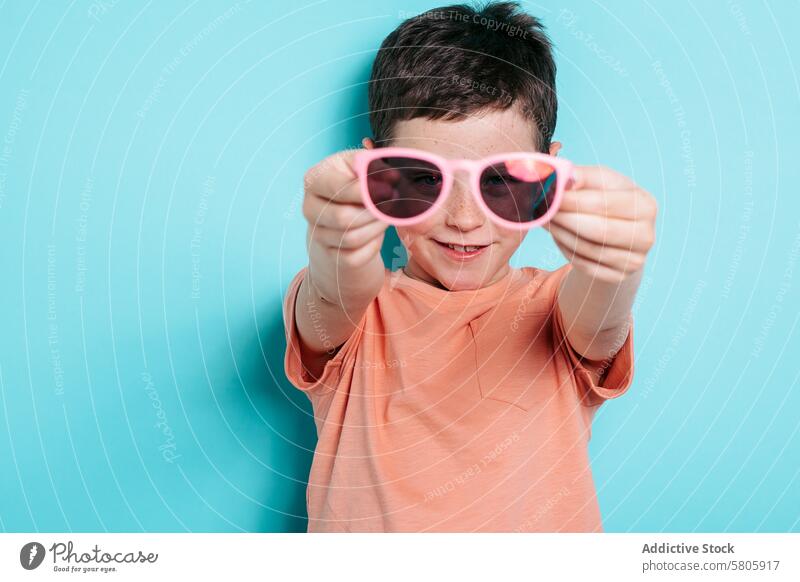 Lächelnder Junge mit rosa Sonnenbrille vor einem blauen Hintergrund Kind Schüler Schule Brille Spaß spielerisch heiter Glück Freude lässig Bekleidung Mode