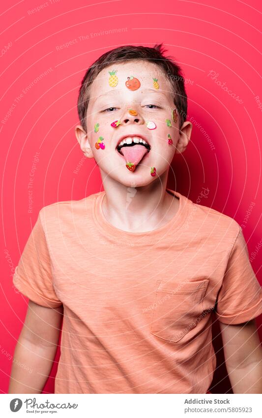 Verspielter Junge mit Sticker-bedecktem Gesicht auf rotem Hintergrund Kind spielerisch Aufkleber Zunge roter Hintergrund Schule in die Kamera schauen heiter
