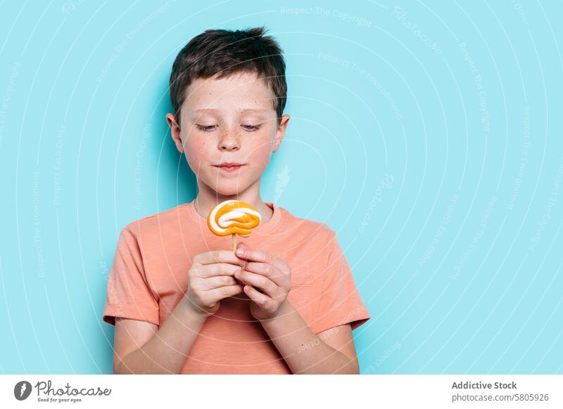 Kleiner Junge bewundert einen Lutscher auf blauem Hintergrund Schulkind Lollipop Leckerbissen Snack süß Bonbon herabsehend Gelassenheit betrachtend Kind