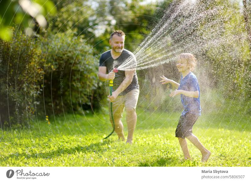Lustiger kleiner Junge mit seinem Vater spielt mit Gartenschlauch in sonnigen Hinterhof. Vorschüler Kind mit Spaß mit Spray von Wasser. Sommer im Freien Aktivität für Kinder.