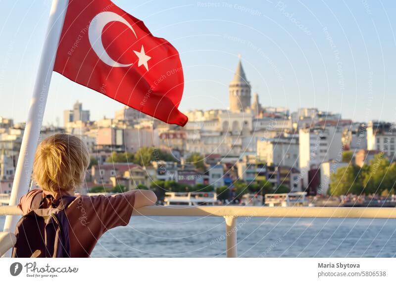 Ein Junge im Teenageralter bewundert vom Ausflugsboot aus den Blick auf das Goldene Horn bei Sonnenuntergang. In der Ferne ist der berühmte Galata-Turm zu sehen. Geschäftiges Stadtbild von Istanbul mit türkischer Flagge im Vordergrund