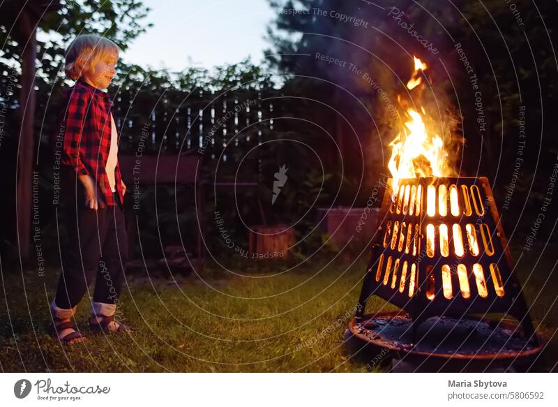 Süßer kleiner Junge brennt ein Lagerfeuer an einem Sommerabend im Hinterhof. Kind legt Brennholz in einer Feuerschale mit Zangen. Sommerferien für Kinder im Dorf.