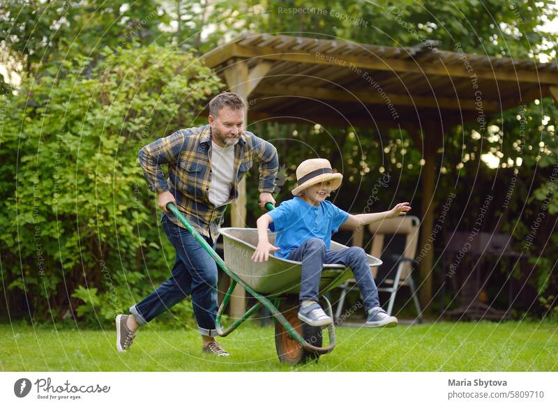 Schubkarre schieben von Papa im heimischen Garten an einem warmen sonnigen Tag. Aktive Spiele im Freien für Familie mit Kindern im Hinterhof Spaß Arbeit Vater