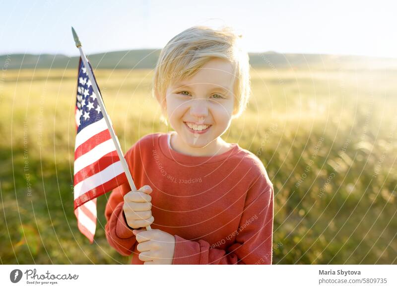 Niedlicher kleiner Junge, der den 4. Juli, den Unabhängigkeitstag der USA, bei sonnigem Sommer-Sonnenuntergang feiert. Kind läuft mit amerikanischer Flagge der Vereinigten Staaten auf Weizenfeld.