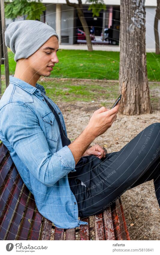 Mann in Jeans und Mütze mit Smartphone auf einer Bank. jung Jeansstoff Jacke Beanie Park fokussiert Bäume Gras lässig im Freien Sitzen Technik & Technologie