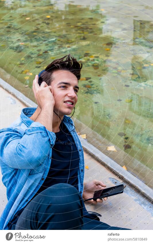 Mann hört Musik an einem Teich. Kopfhörer Smartphone hören Jeansstoff Hemd schwarz Hose jung Erwachsener fokussiert entspannt urban im Freien Sitzen lässig
