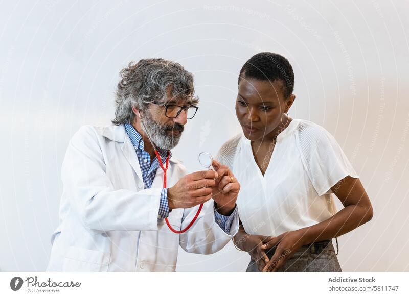Älterer Arzt zeigt einem Patienten sein Stethoskop zeigend geduldig prüfen Ernennung untersuchen Krankenhaus Gesundheitswesen besuchen multiethnisch