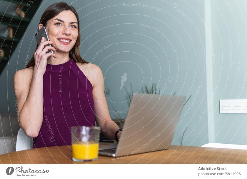 Lächelnde Frau, die ein Smartphone benutzt und mit einem Laptop am Tisch sitzt benutzend Zahnfarbenes Lächeln Apparatur Browsen Telefon Inhalt heiter Netbook