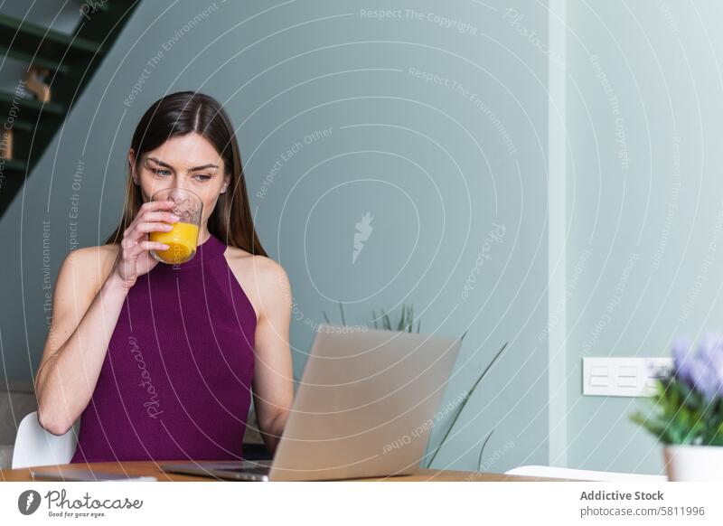 Fröhliche Frau, die im Wohnzimmer am Laptop sitzt benutzend Zahnfarbenes Lächeln Glück Apparatur Browsen Saft trinken heiter Netbook lässig Glas orange