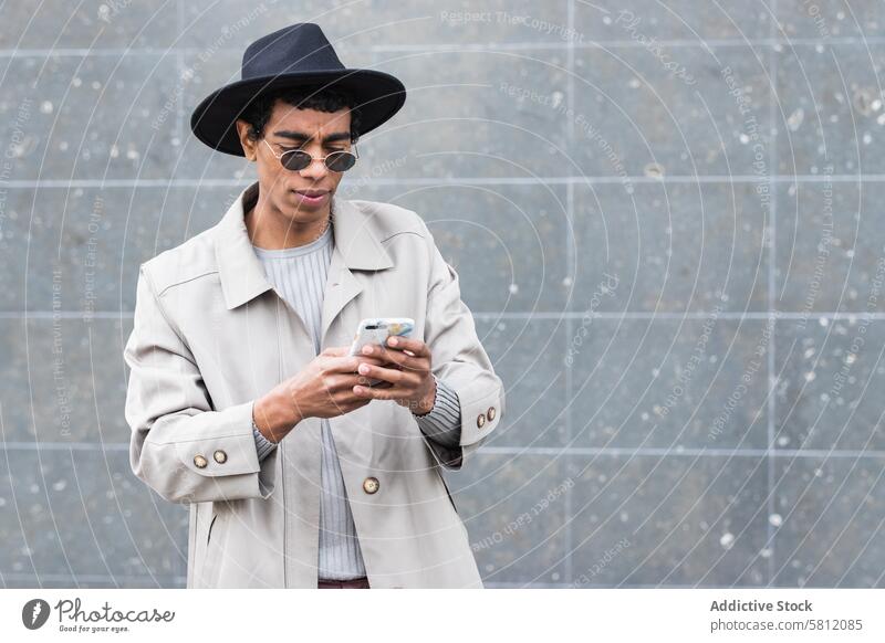 Stilvoller ethnischer Mann mit Smartphone auf der Straße genießen trendy Drahtlos benutzend Apparatur Gerät Internet online Funktelefon Hut Kleidungsstück Mode