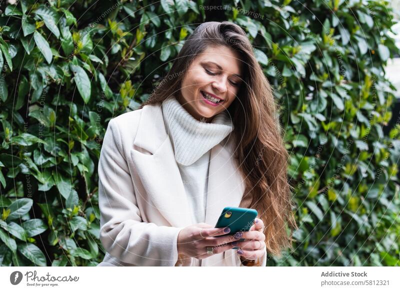 Zufriedene Frau benutzt Smartphone im Park benutzend Glück Stil Telefon heiter Mobile Browsen Kommunizieren positiv Erwachsener elegant Oberbekleidung Mantel
