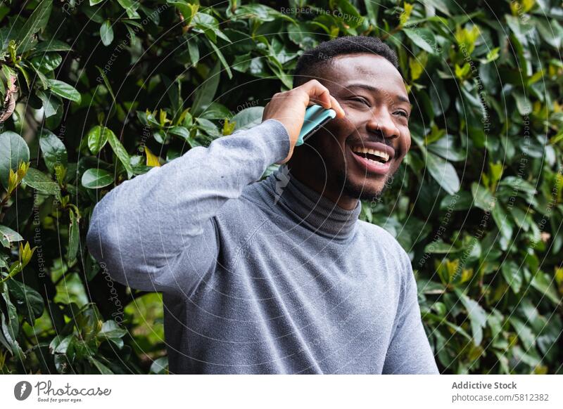 Fröhlicher schwarzer Mann, der im Park mit seinem Smartphone spricht reden Glück Telefon Gespräch heiter Lachen Mobile männlich jung Afroamerikaner ethnisch
