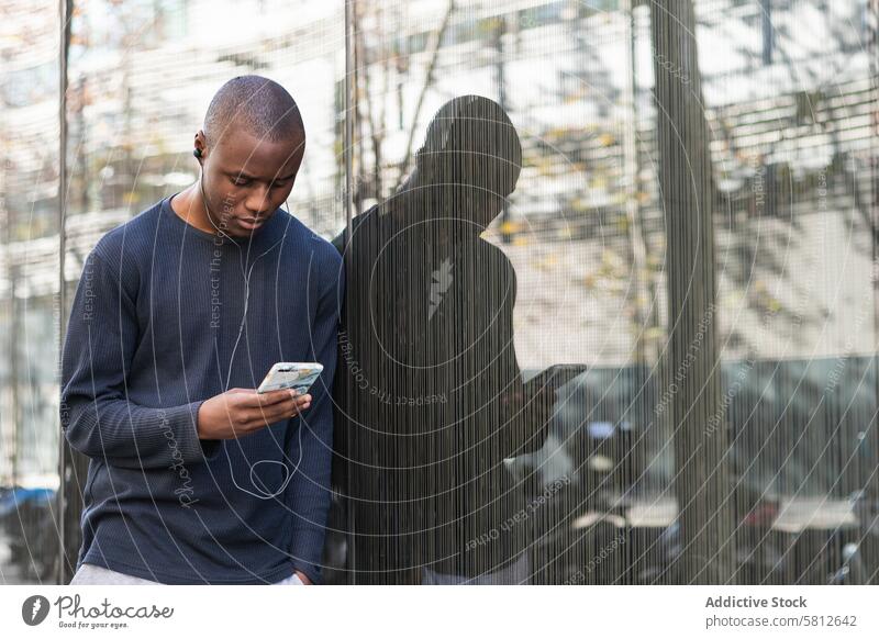 Seriöser schwarzer Mann mit Smartphone vor grauer Wand benutzend ernst Kopfhörer Textnachricht achtsam Musik zuschauen prüfen lesen digital männlich Mobile