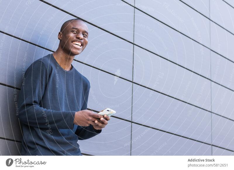 Glücklicher schwarzer Mann mit Smartphone vor grauer Wand benutzend Textnachricht Lächeln zuschauen prüfen lesen digital männlich Mobile Surfen Telefon