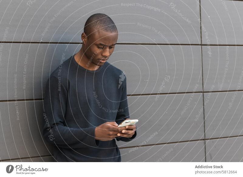 Seriöser schwarzer Mann mit Smartphone vor grauer Wand benutzend ernst Textnachricht achtsam zuschauen prüfen lesen digital männlich Mobile Surfen Telefon