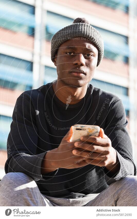 Ernster schwarzer Mann mit Smartphone auf der Straße sitzend benutzend ernst Fokus Browsen Anschluss Denken Internet digital männlich online jung Funktelefon
