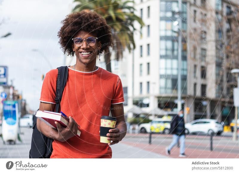 Fröhlicher junger Schwarzer auf der Straße mit Kaffee zum Mitnehmen und Lehrbüchern in der Hand Mann Spaziergang Lächeln Schüler Buch Imbissbude Großstadt Glück