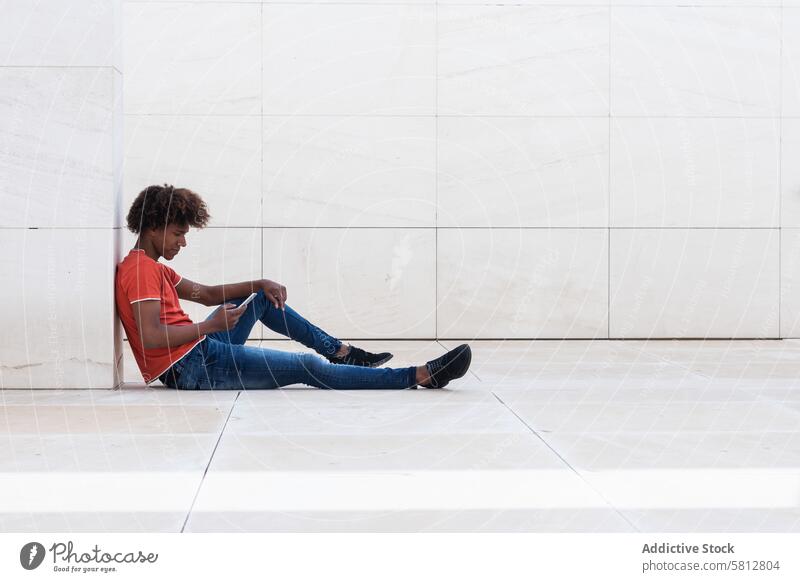 Konzentrierter junger ethnischer Mann, der auf dem Boden sitzt und ein Smartphone benutzt benutzend ernst SMS digital Nachricht Talkrunde Fokus online Mobile
