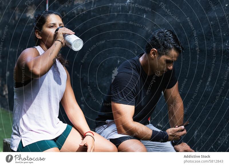 Ethnische Frau trinkt Wasser in der Nähe eines Sportlers mit Smartphone Mann Athlet Training benutzend Durst trinken Pause Sportlerin Paar Internet Funktelefon
