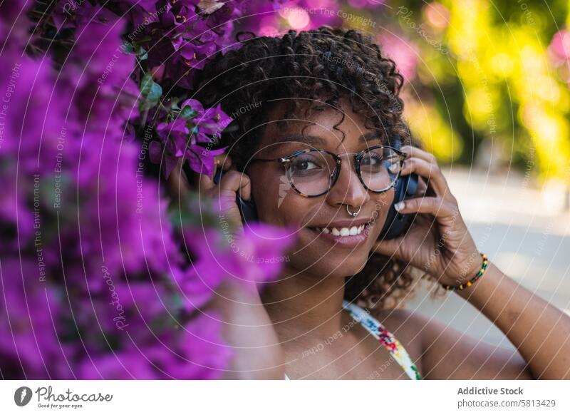 Schwarze Frau hört Musik in der Nähe von Blumen zuhören Buchse Garten Kopfhörer Lächeln Stil jung Brille Piercing ethnisch schwarz Afroamerikaner Gesang Freude