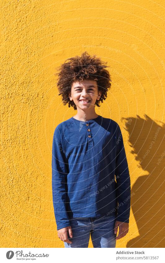 Ethnischer Junge mit lockigem Haar an gelber Wand stehend Afro-Look krause Haare farbenfroh hell Kind Teenager gestikulieren positiv männlich Behaarung