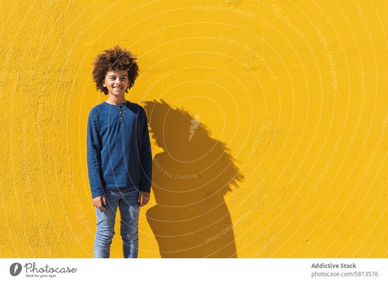Ethnischer Junge mit lockigem Haar an gelber Wand stehend Afro-Look krause Haare farbenfroh hell Kind Teenager gestikulieren positiv männlich Behaarung