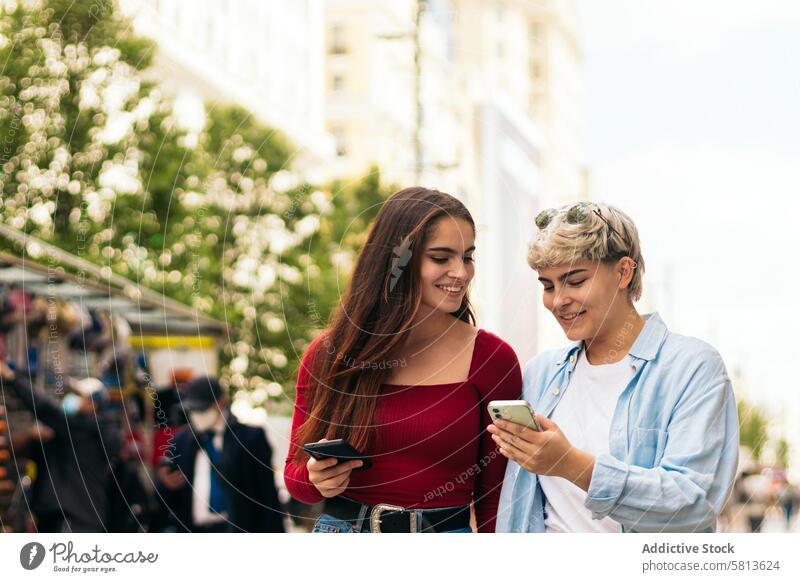 zwei Teenager-Mädchen, die spazieren gehen und das Handy beobachten Telefon Mobile Blick Smartphone zuschauend Beteiligung Lachen Lächeln Internet App im Freien