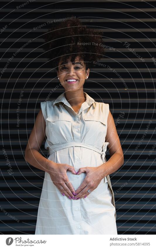 Lächelnde schwarze Frau zeigt Herzgeste auf schwangeren Bauch gestikulieren mütterlich Glück erwarten vorwegnehmen pränatal positiv Schwangerschaft froh