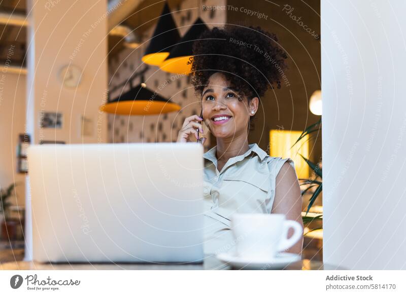 Lächelnde schwarze Frau, die an einem Tisch mit Laptop über ihr Smartphone spricht reden benutzend positiv sprechen Telefonanruf Glück Anschluss freiberuflich