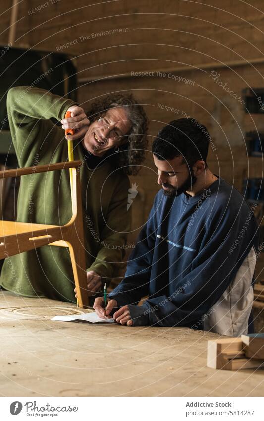 Handwerker, der einen Holzstuhl herstellt, während ein Kollege Skizzen zeichnet Männer Tischlerin Mitarbeiterin messen Klebeband Stuhl Werkstatt Tischlerarbeit