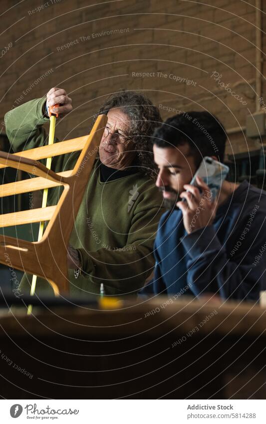Konzentrierter Mann spricht mit seinem Smartphone und schreibt Notizen in einer Schreinerei Männer Zimmerer Kollege Klebeband messen Stuhl benutzend Orden