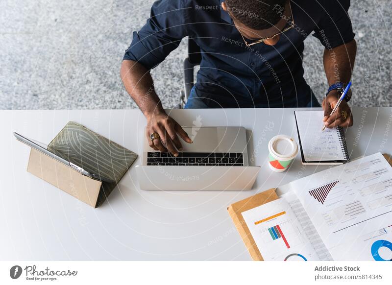 Afroamerikanischer Mann, der in der Nähe eines Laptops im Büro in einen Notizblock schreibt zur Kenntnis nehmen Notebook Netbook Tisch Schriftstück Mitarbeiter