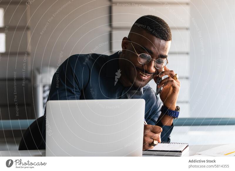 Schwarzer Mann telefoniert in der Nähe eines Laptops im Büro Telefonanruf zur Kenntnis nehmen Notizblock Smartphone Lächeln Tisch Schriftstück Afroamerikaner