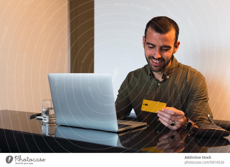 Fröhlicher Mann mit Kreditkarte beim Online-Shopping Laptop online Zahlung Transaktion kaufen Kauf Finanzen e-Commerce abgelegen Netbook männlich Tisch Gerät