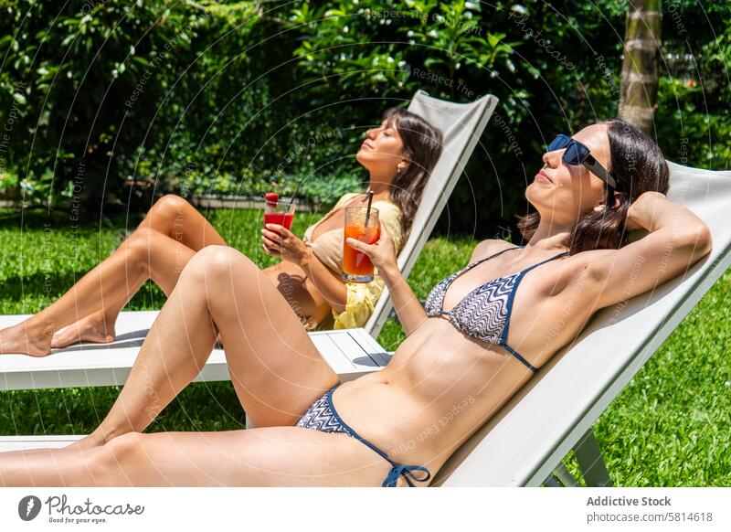 Zwei Freunde sonnen sich auf Liegestühlen mit Fruchtsmoothies in der Hand attraktiv schön beste Getränk Bikini Bonden sorgenfrei Stuhl Hof Liegestuhl trinken