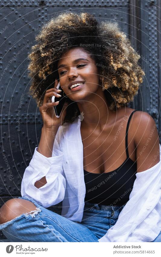 Entzückte schwarze Frau, die mit ihrem Smartphone spricht, während sie in der Nähe eines Gebäudes sitzt positiv Telefonanruf sprechen benutzend Kommunizieren